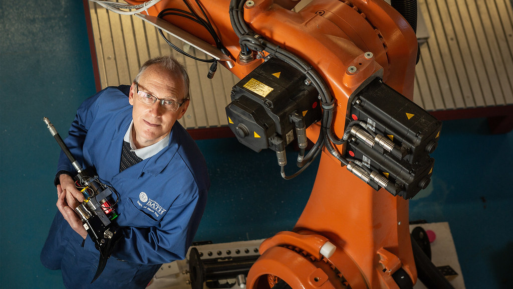 Professor Andrew Plummer holds a hydraulic ankle prototype next to a robotic arm.