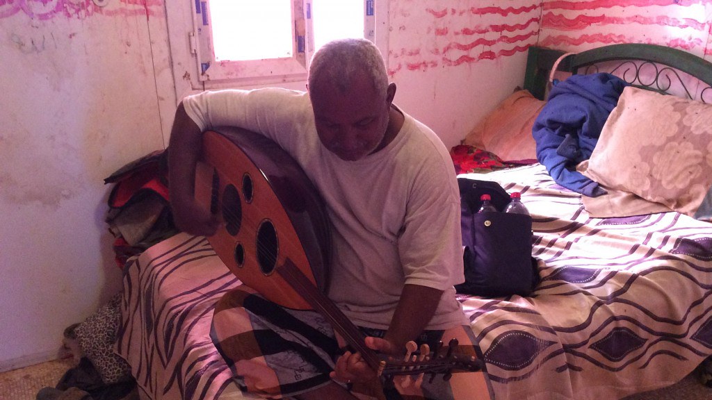 在一间破旧的小房间里，一个黑人坐在床上弹奏乌德琴 