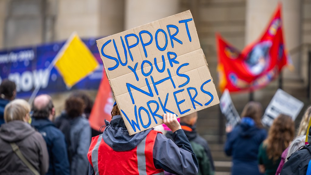 A photo showing a striking worker carrying a banner which says 'Support your NHS Workers'