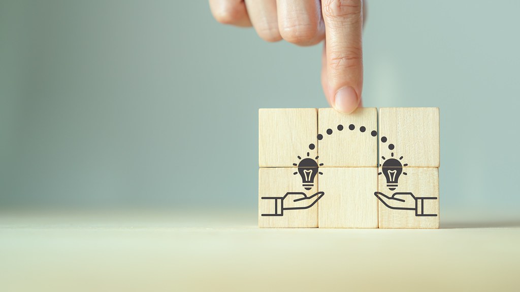 A photo of a finger touching building blocks marked with an image of two hands holding lightbulbs and transferring ideas