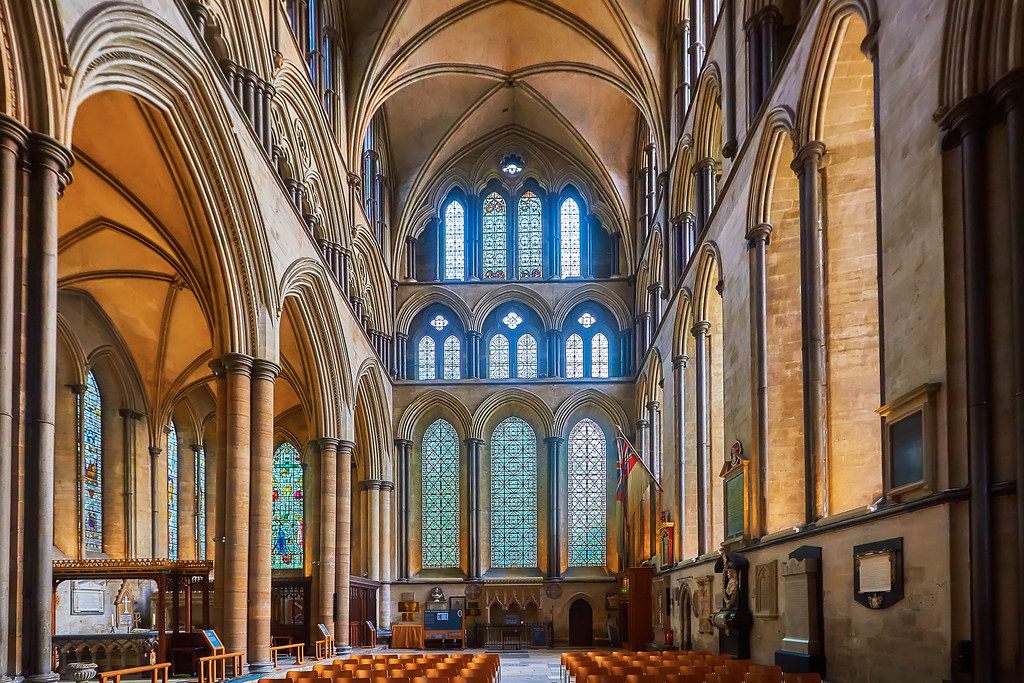 East end of a cathedral in Salisbury, England. The wall is covered in ornate arched windows separated into 3 distinct rows. Moreover arches can be seen on either side of the windows throughout the cathedral. 
