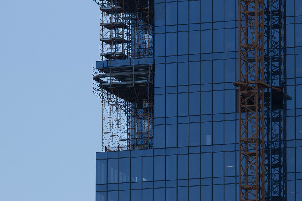 A concrete frame rising above a city street, with formwork on the upper slab and a tower crane standing beside the unfinished structure.