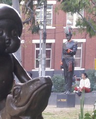 Near us, a statue of a baby wearing a gumnut hat riding on the back of a lizard. Further away, a statue of Victoria's Lieutenant-Governor from the 1850s, Charles La Trobe, in front of someone sitting cross-legged.   State Library forecourt, Swanston Stree