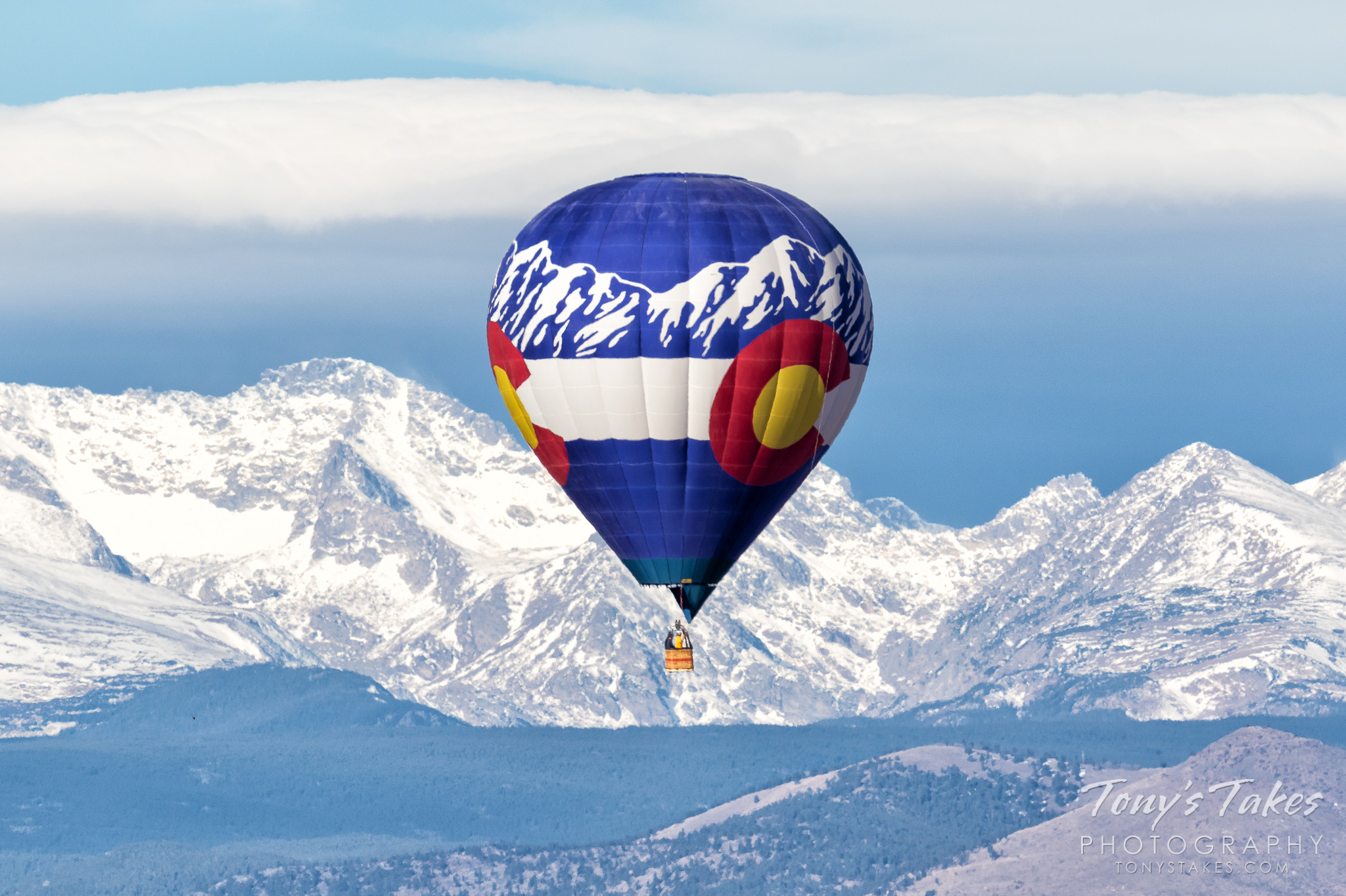 A hot air balloon looking like a Colorado flag flies in front of the Rocky Mountains. (© Tony’s Takes)