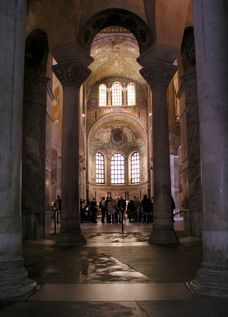 Inside of a large domed building. There are two columns in the front of a domed-area with tourists and larged arched windows. Moreover, the dome is covered in intricate religious paintings. 