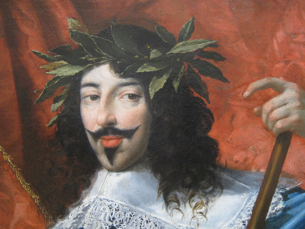 Painting of Louis XIII of France. He wears a plant-crown, is in a white outfit, has pale skin and dark hair. Moreover, he is standing in front of a dark red background. 