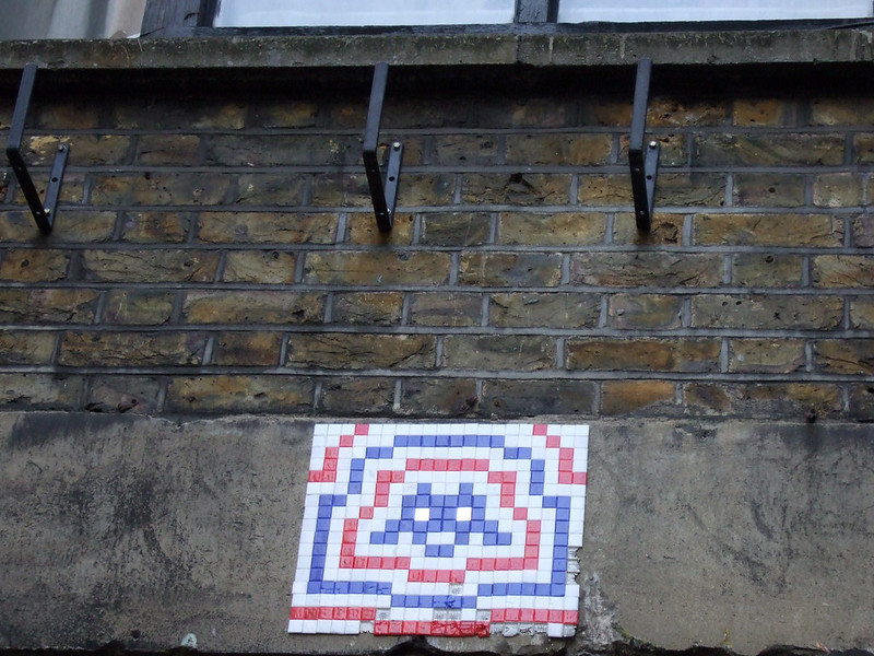 Brown and beige brick wall with a cement base decorated iwth white, blue, and red square tiles that look like an alien from a vintage video game