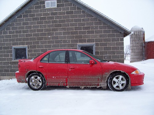 Side view of red car in front of brick building in the snow, with ice and snow grime on it
