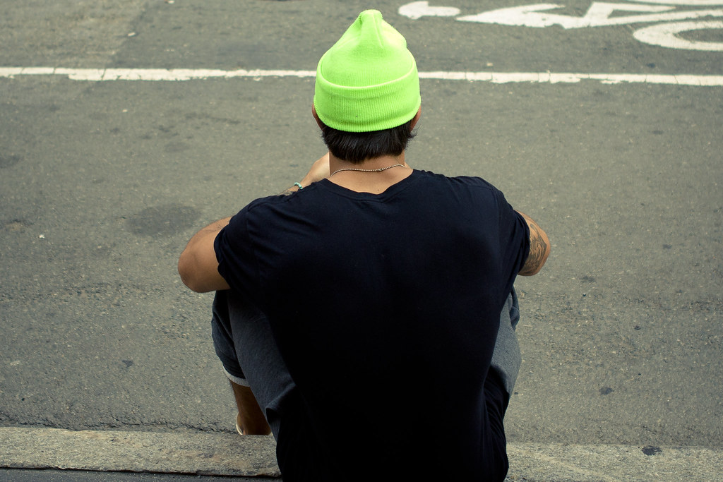 Young man wearing a neon green beanie and black t-shirt sitting on a New York City street curb.