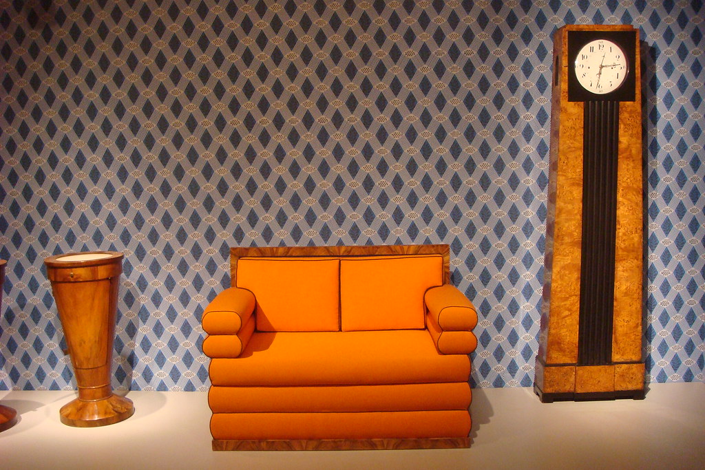 Biedermeier pieces: An orange couch in the center, with a medium wood clock and a cone-shaped table on the right and left, respectively. 