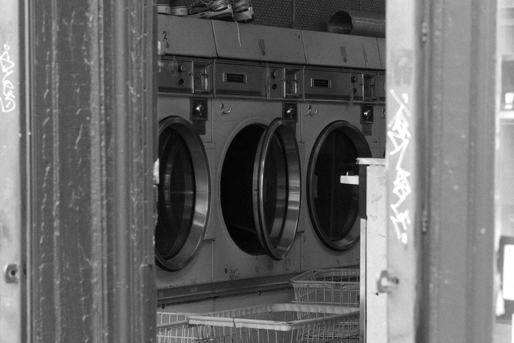 Open doorway into an NYC laundromat showing rows of industrial dryers and rolling wire carts.