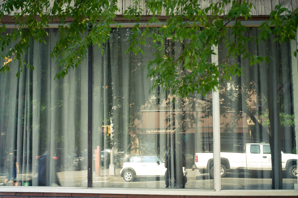 Storefront window with sheer curtains reflecting trees, a white pickup truck, and a Mini Cooper.