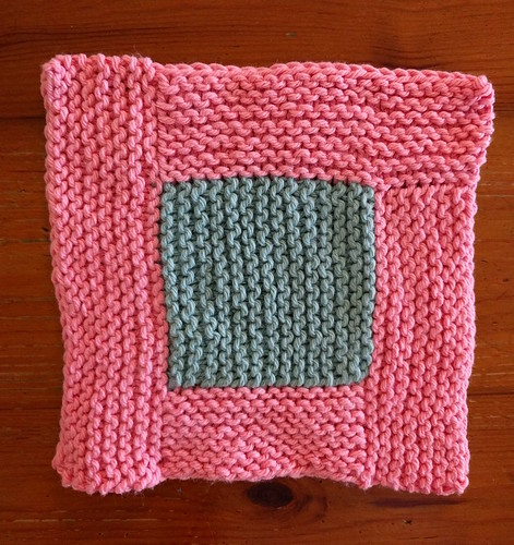 A dishcloth made in a garter stitch log cabin style, with rectangular pink borders surrounding a blue square centre.