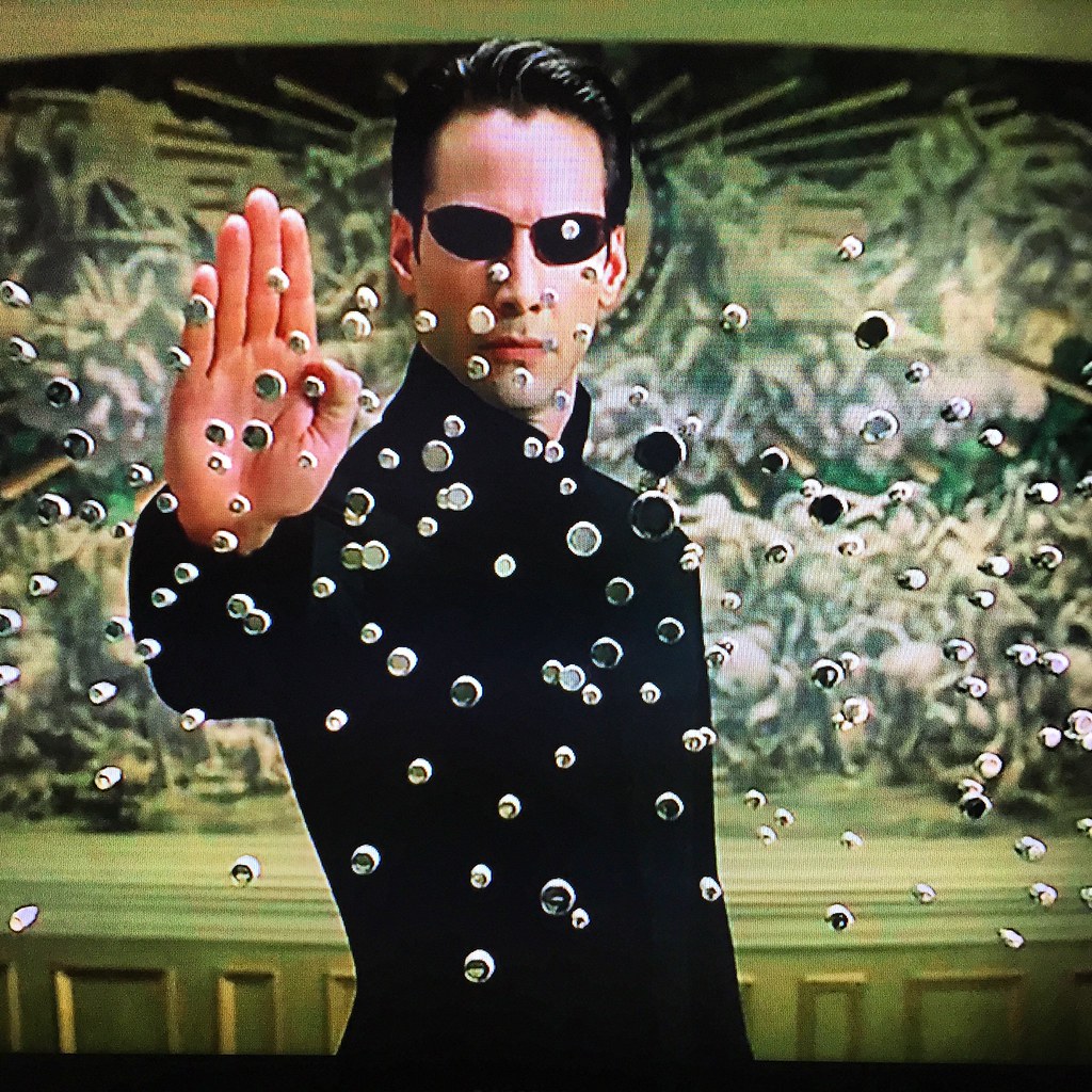 A still frame from the movie _The Matrix Reloaded (2003), from the scene where Neo stops all the gunshots from The Merovingian's henchmen by merely raising his hand in a "stop" sign. The picture seems pixelated, and there seems to be a curve to it, which suggests this picture was taken with a smartphone camera while the movie was playing on the TV.