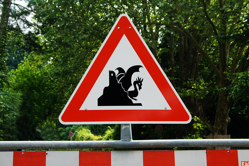A road sign depicting a dragon sitting on the side of a mountain beside a ruined tower.
