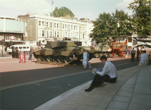 MAN SITTING ON THE STEPS READING OUTSIDE EARLS COURT ENTERTAINMENT AND EXHIBITION CENTRE WITH A MILITARY TANK AND PEOPLE WALKING AND STANDING IN LONDON ENGLAND   1997 a . THIS IS A WELL KNOWN VENUE IN THE GB WHICH I THINK HAS BEEN OR IS GOING TO BE DEMOLI