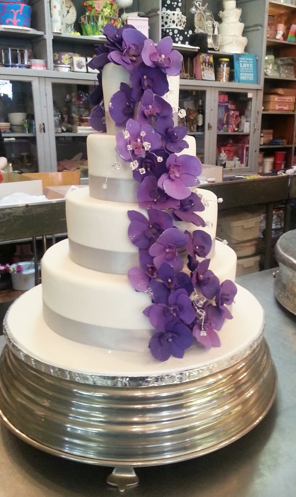 4-tier Wicked Chocolate wedding cake covered in white fondant icing, decorated with cascade of purple fondant orchids with sprigs of non-edible crystals & silver satin ribbon at bases