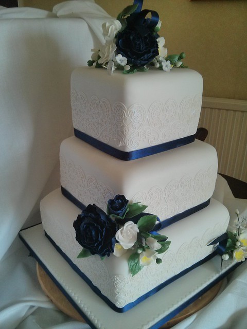 Three tier stacked wedding cake. Claire Bowman lace borders (tastes like marshmallow, very easy to work with). Matching flower  sprays, handcrafted from sugar paste. Colour themed navy blue ribbons. Navy blue  flower paste peony flowers with white roses.
