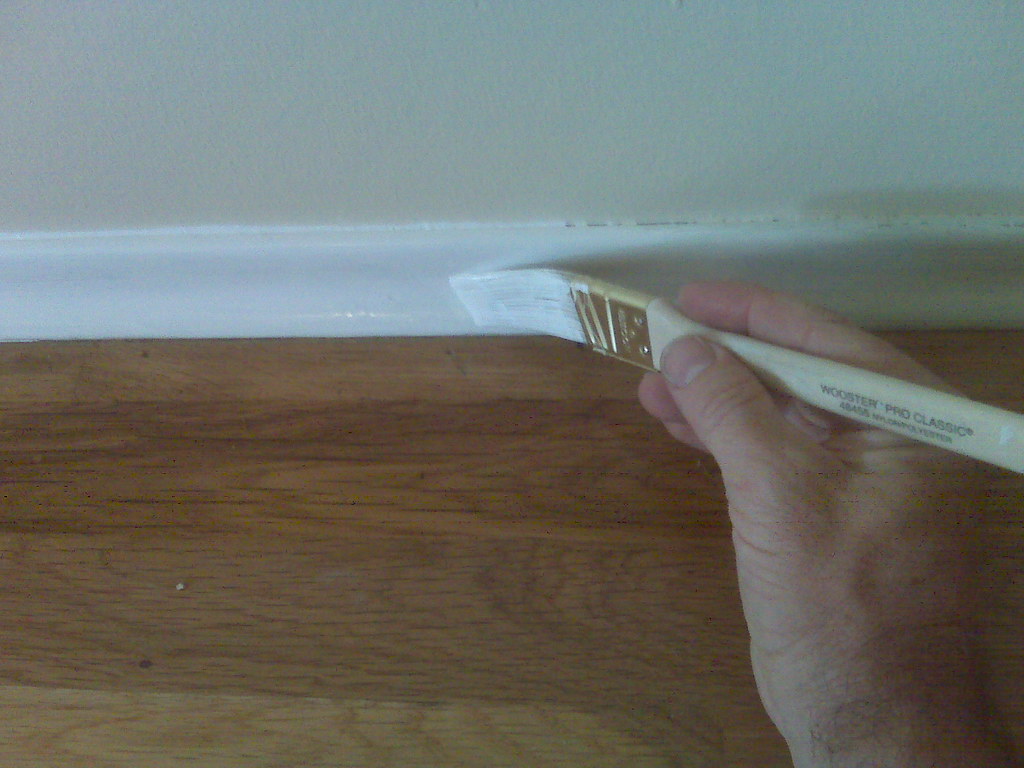 Brush laying white enamel along a bedroom baseboard above a hardwood floor