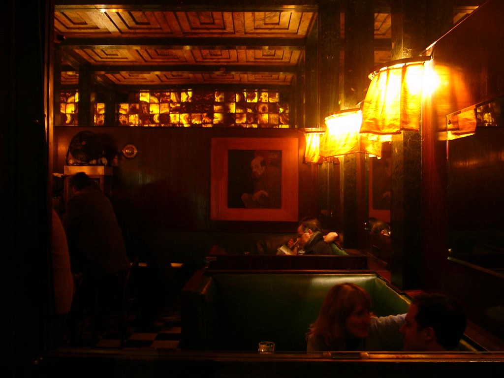 American Bar, Vienne, Autriche, 1907, Adolf Loos : Un bar sombre éclairé avec des lampes aux tons chauds le long du mur droit et des éléments simples et romantiques dans tout l'espace.
