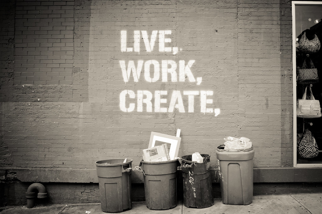 Painted brick wall with stenciled text reading LIVE, WORK, CREATE above four curbside trash cans.