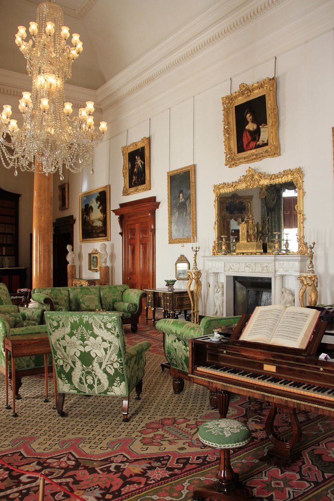 A room decorated in the Regency style. There is an ornate chandelier in the center, various gold-framed portraits, green floral furniture, a white fireplace and a medium-wood piano.