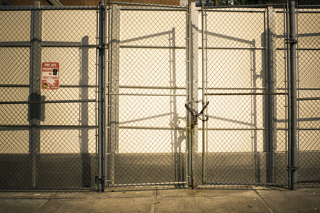 Closed chain-link gate padlocked before a white security wall with geometric afternoon shadows.