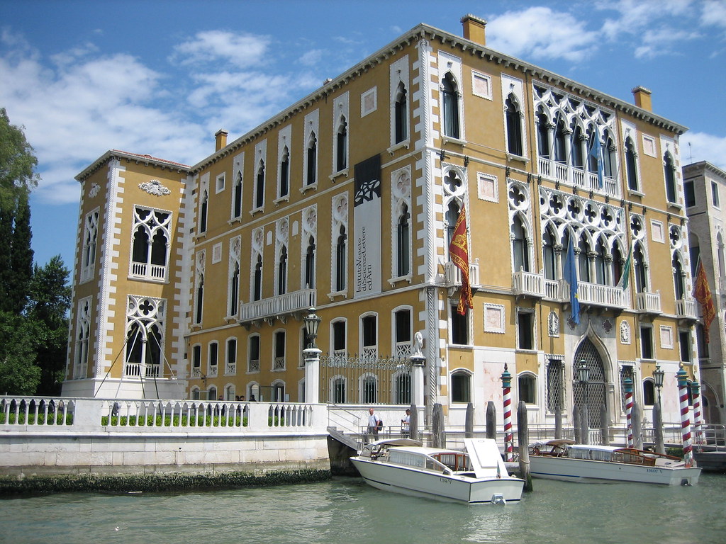 A photo of the Palazzo Cavalli Franchetti, which is a large rectangular structure that sits right along the grand canal in Venice. Moreover, this structure is a mustard color with white arched windows and various decorations. 