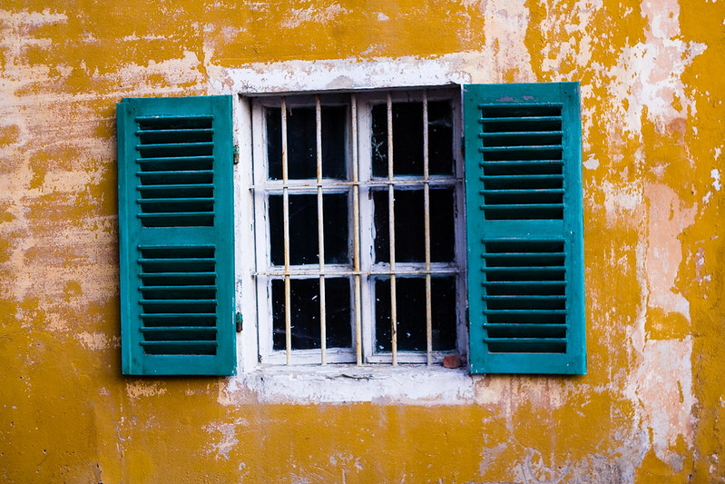 Yellow and white exterior wall with a white-framed window with turquoise shutters open on both sides