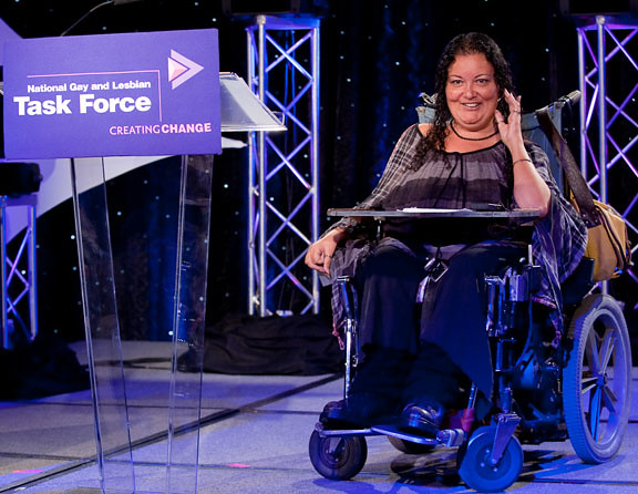 A photo of Patricia Berne at the National Gay and Lesbian Task Force conference. She is in an active speaking pose with her hand up and her mouth smiling. She sits in her mobility aid. 