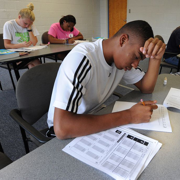 A focused student sits at his desk, taking a test, while two other students behind him are also engaged in their tests.