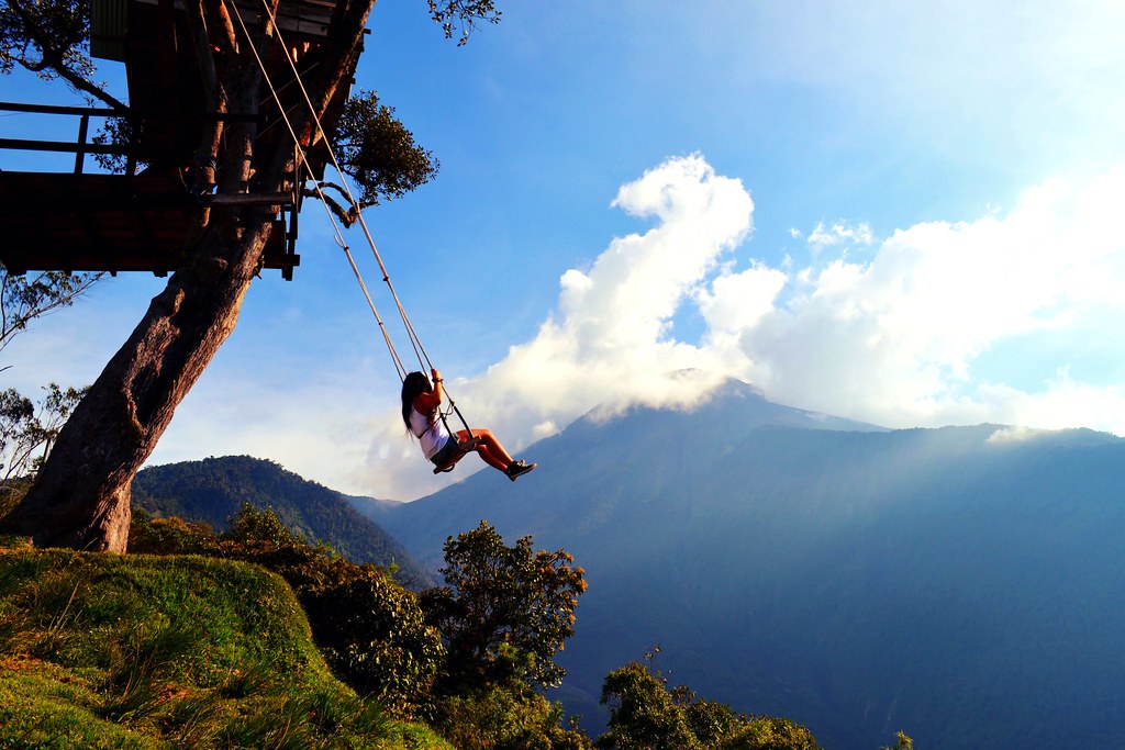 A professional image of a woman swinging on a swing at La casa del árbol in Equator. The mountains in the background are visible against a sunny blue sky with light clouds.