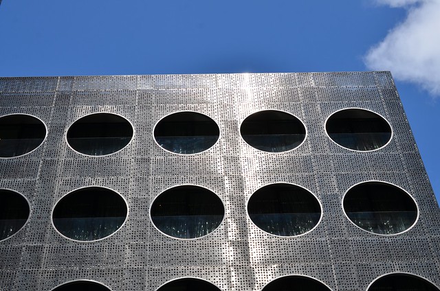Perforated metal facade with evenly spaced circular openings in a grid against a bright blue sky.