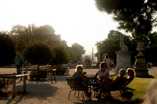An image of the Tuileries Gardens in Paris. Trees line a wide boulevard and people sit outside on metal chairs.