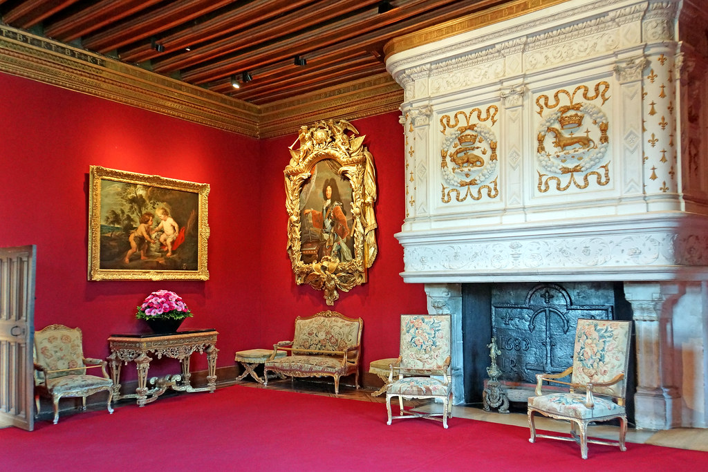 Louis XIV's Drawing Room with deep red walls, gold picture frames, a large white fireplace and light-colored chairs sit around the room. 