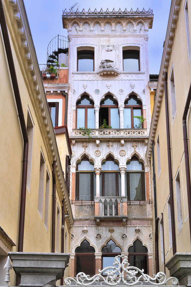 Photo of an alley that has a tall skinny building at the end that has three levels of Gothic windows. Located in Venice Italy 
