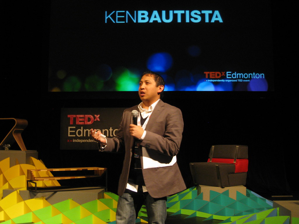 Ken Bautista speaks into a microphone in front of signs for TEDxEdmonton