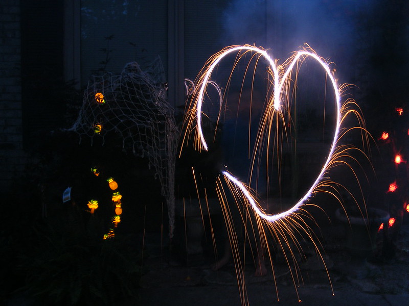 dark background with bits of light around a heart drawn with a sparkler, amber light shooting off the white heart.