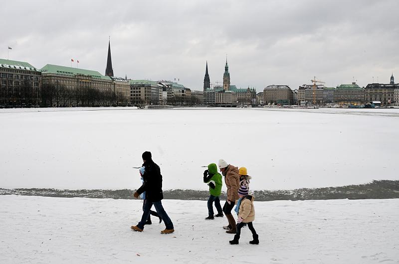 4983 Winter in Hamburg, die Binnenalster ist mit Eis und Schnee bedeckt - eine Familie gehtm am Alsterufer spazieren - im Hintergrund die Hamburger Altstadt.