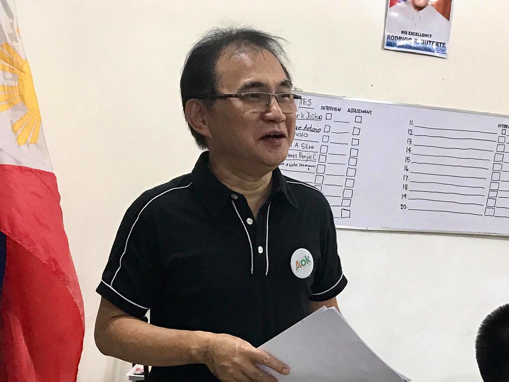 The image shows Mr. Uswaldo Parrenas wearing black shirt and eyeglasses. He speaks about Autism Works and holds papers.