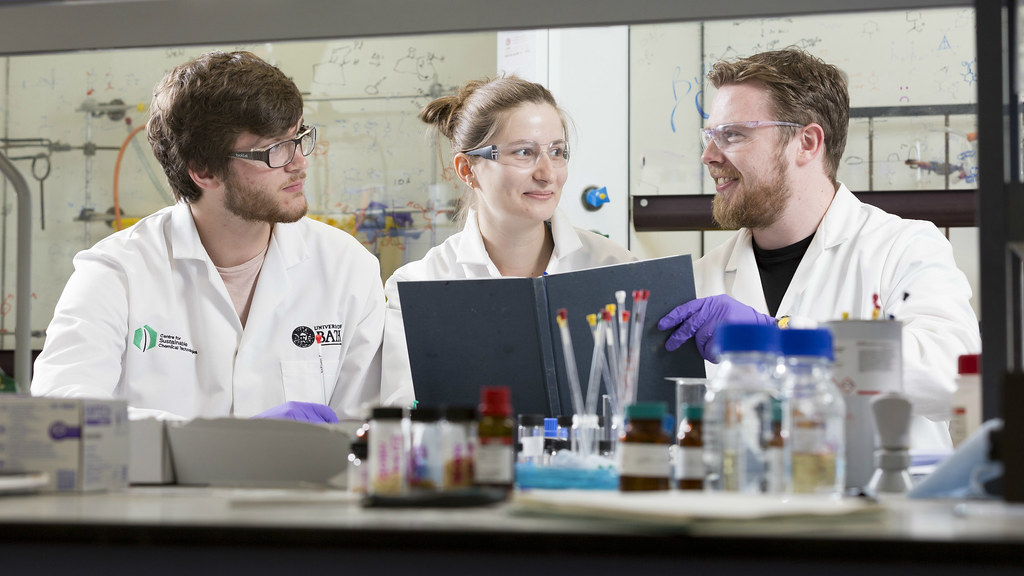 3 PhD students , wearing white lab coats, sitting in a laboratory, looking at a notebook