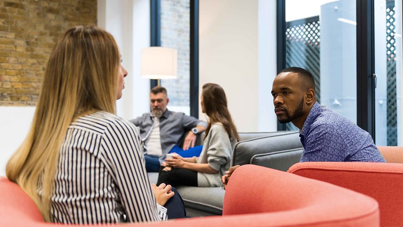 Two people sit in armchairs talking intently to one another. In the background, another two people do the same.  