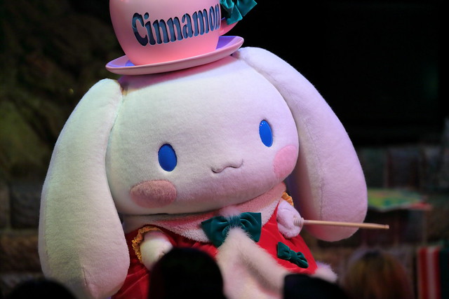 Cinnamon bunny mascot on stage with pink hat labeled Cinnamon, cream fur, blue eyes, rosy cheeks, red dress with teal bows, holding a wand; audience silhouettes in foreground.
