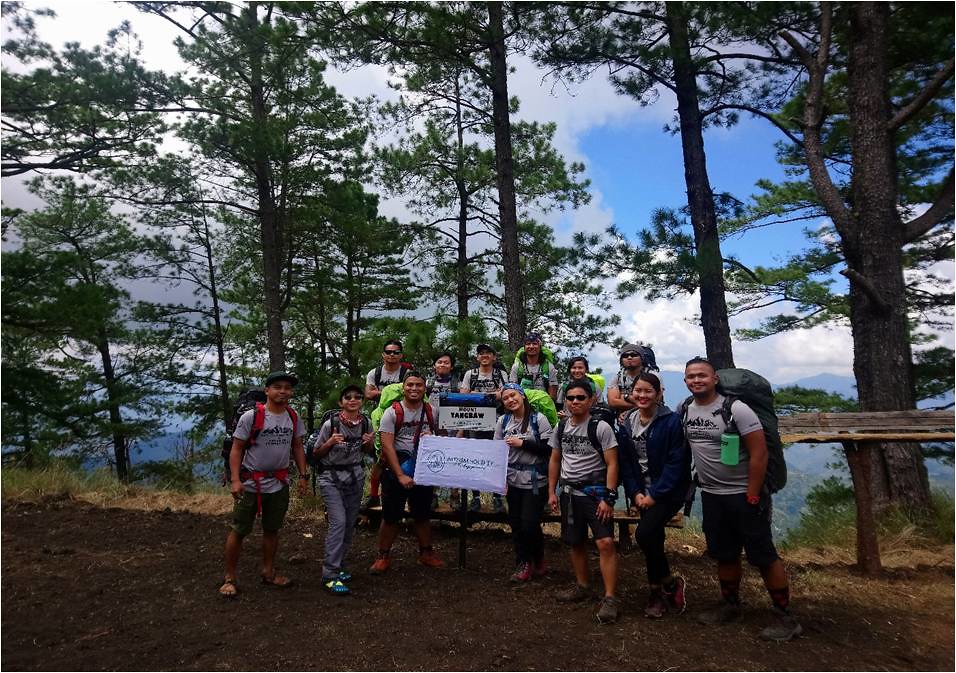 The image shows Hidden Leaf Mountaineers and Burdados Mountaineering Club at the peak of Mt. Tangbaw, Bokod Benguet standing while having a group photo with a view of lots of pine trees.