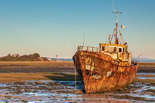 A rusty abandoned fishing vessel beached off the causeway to Roa Island. Taken with the glow of sunset.  Rampside Lighthouse (“The Needle”) is shown in the background.  Photo by John Roberts.
