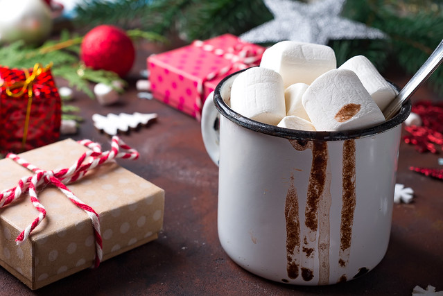 Enamel cup of hot cocoa with mini marshmallows with pine boughs against a white background with beautiful Christmas gift box. Close up