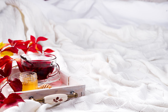 Hot healthy rose tea with honey on wooden tray with autumn fallen leaves on knitted warm woolen blanket. Relaxing in cold weather on the bed with copy space . Seasonal beverages.