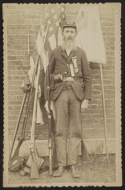 [Unidentified Civil War veteran from Grand Army of the Republic post 386 in uniform with musket in front of flags, weapons, and equipment] (LOC)