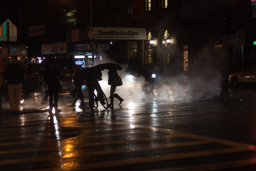 Silhouetted pedestrians with umbrellas and a cyclist on rain-slicked asphalt outside DavidBartonGym.