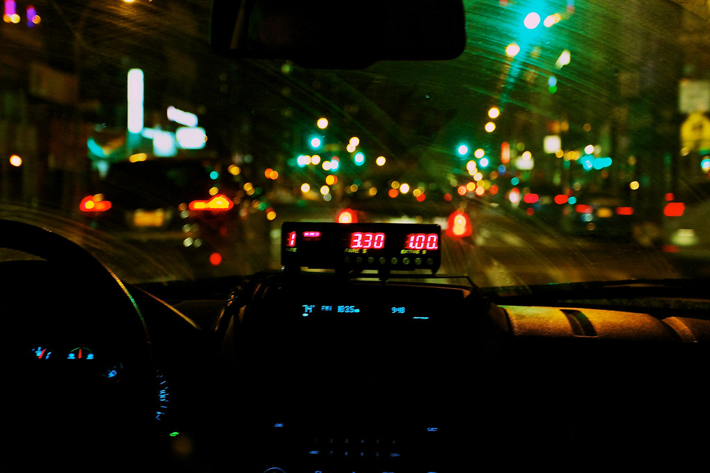 Taxi windshield view of shimmering bokeh streetlights with a red fare meter on the dashboard.